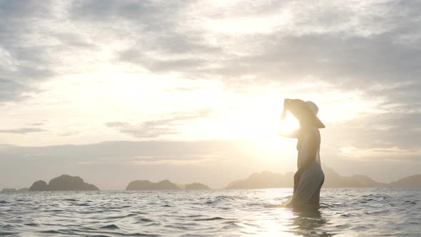 Woman In Bikini And Sunhat Standing In Sea As Sun Sets