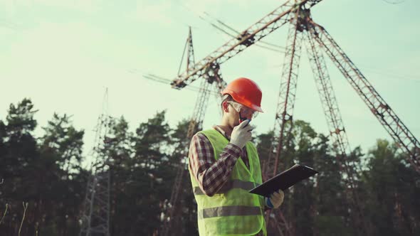 Caucasian Engineer in Protective Uniform and Orange Hard Hat Holds Walkietalkie and Clipboard on