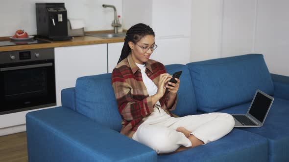 Young Woman Using Smartphone at Home on a Couch with Laptop
