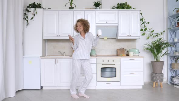 Young Excited Woman Dancing In Pajamas Holding Smartphone In Kitchen