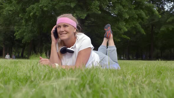 Smiling Senior Woman Talking on Smartphone Relaxing on Grass After Workout in Park