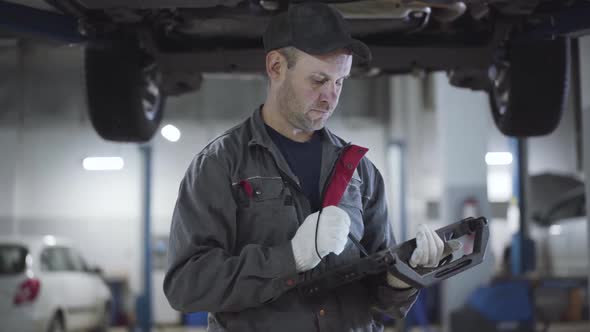 Confident Adult Maintenance Engineer Standing Under Car Bottom in Repair Shop and Using Computer