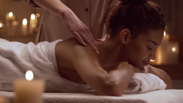 Young Tranquil African American Female Lying on Table in Day Spa and Enjoying Massage of Neck