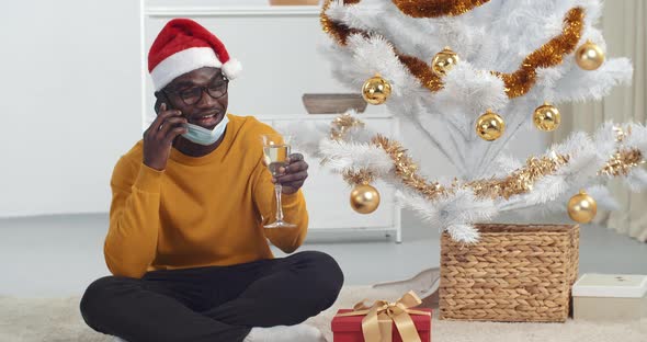 Afro American Guy Black Man Sitting Under Christmas Tree Wears Santa Hat and Medical Protective Mask