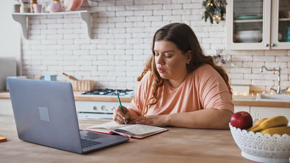 Young Overweight Woman Watching Webinar on Laptop and Making Notes Sitting in Kitchen at Home