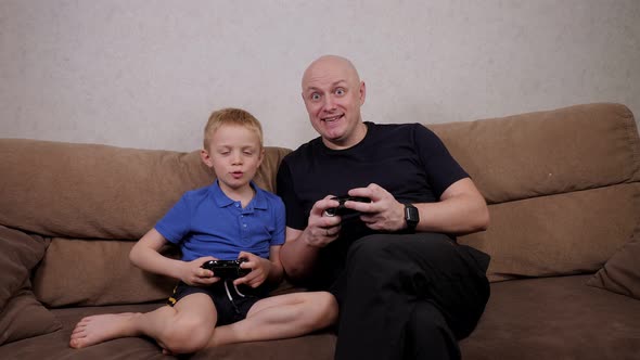 Father and Son Play Video Games on Consoles at Home on the Sofa