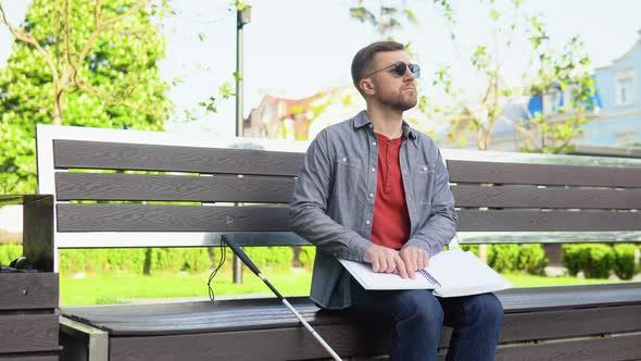 Young Blind Man Sitting on Bench in City Park and Reading a Braille Book