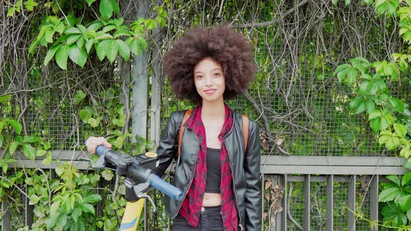 Young woman standing in front of overgrown fence, holding e-scooter