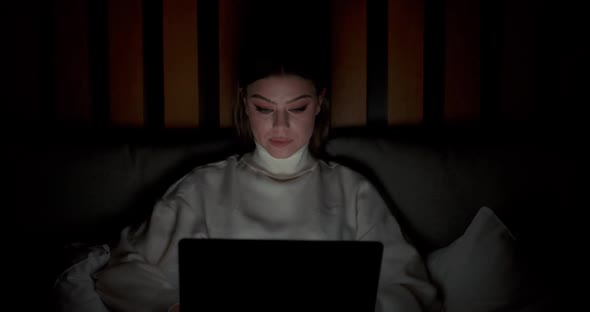 Young Woman Lying on the Bed and Working on a Laptop Types Text on the Keyboard Night Time Remote