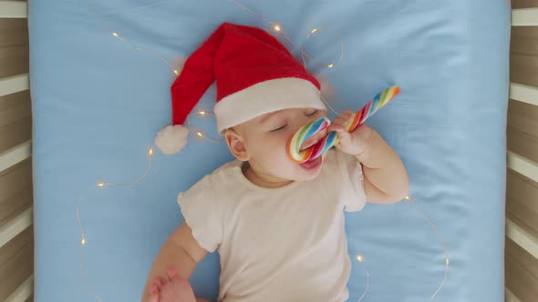 Smiling Funny Newborn Baby in Santa Claus Hat Red Bodysuit Play on White Soft Bed Lick Sweet