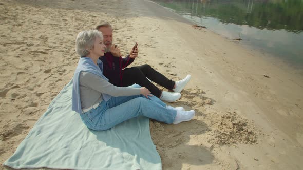 Mature Couple Sitting on Sand Listening to Music Via Smartphone