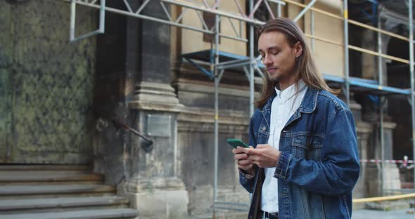 Side View of Long Haired Young Man Typing Message and Touching Smartphone Screen While Walking at