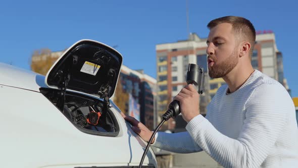 A Lightskinned Male Driver Holds a Charging Cable for an Electric Car Sitting Near an Open Car