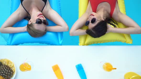 Closeup of Two Stylish Women Relaxing on Inflatable Mattress Next to Pool Bar