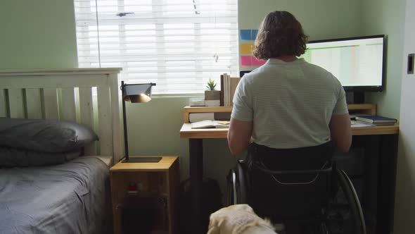 Focused caucasian disabled man in wheelchair using computer in bedroom with his pet dog