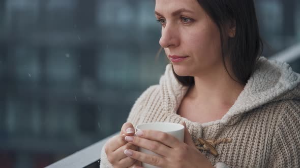 Caucasian Woman Stays on Balcony During Snowfall with Cup of Hot Coffee or Tea