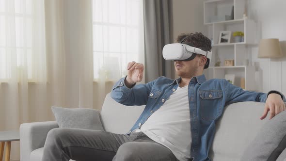 Amazed Guy Watches Online Video to VR and Moves Hands Sitting Against Window on Sofa at Home Slow