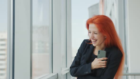 Woman Looking at Camera and Laughing. Red-haired Girl Stands By the Window in the Office and Laughs