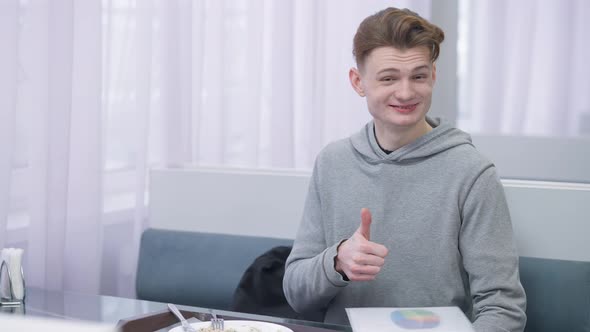 Portrait of Cheerful Millennial Male Student Showing Thumb Up Smiling Looking at Camera Sitting in