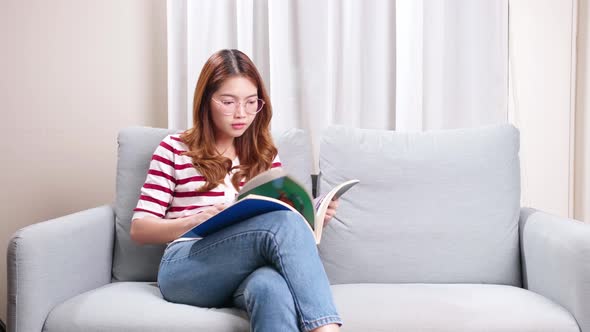 Beautiful young Asian model flipping through picture magazine sitting on sofa at home.