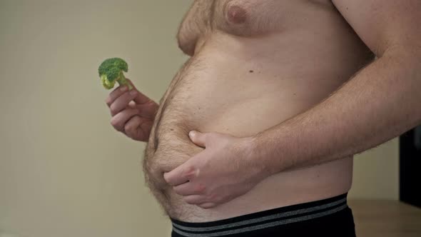 Fat Man Shows the Folds of Excess Fat on His Stomach with One Hand and Holds Broccoli with the Other