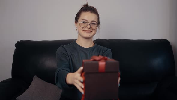 The Portrait of a Redhaired Young Woman with Glasses Stretching the Gift Box Forward Displays a