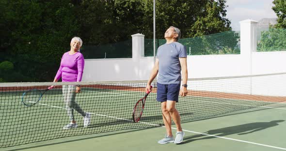 Video of happy biracial senior couple walking with rackets on tennis court