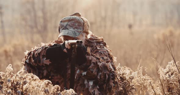 Portrait of Hunter in Hunting Equipment Aims with Shot Gun, Lies in Wait in Field in Sunset Light