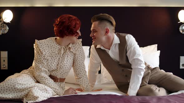 Couple of Man and Woman in Costumes Sit on Bed in Hotel or Apartment and Chat Looking Into Their