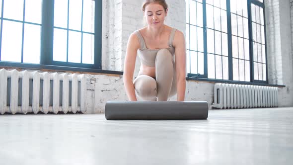 Smiling blonde woman with bun hairstyle in beige tracksuit unrolls mat to camera