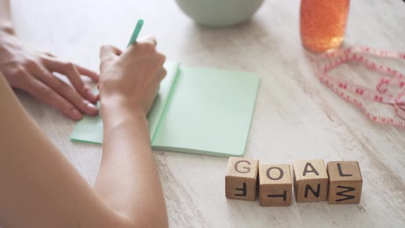 Girl At The Table And Writes In Notebook. Young Woman Sets Goal To Lose Weight. Word Goal On Table