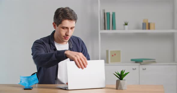 Man Freelancer Sits Down at Table Takes Off His Medical Mask Prepares to Start Work Typing on Laptop