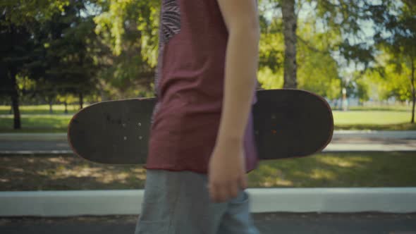 Skateboarder Walks in a Park with a Skateboard in Hands