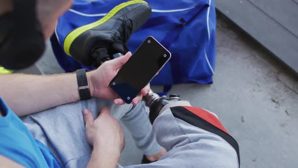 Caucasian disabled male athlete with prosthetic leg sitting, wearing headphones, using smartphone