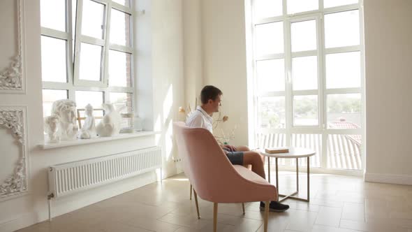 A man is sitting on a sofa in a spacious room, takes a book from a coffee table and begins to read.