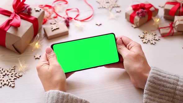 Woman holding smartphone with horizontal green screen on Christmas background with gifts.