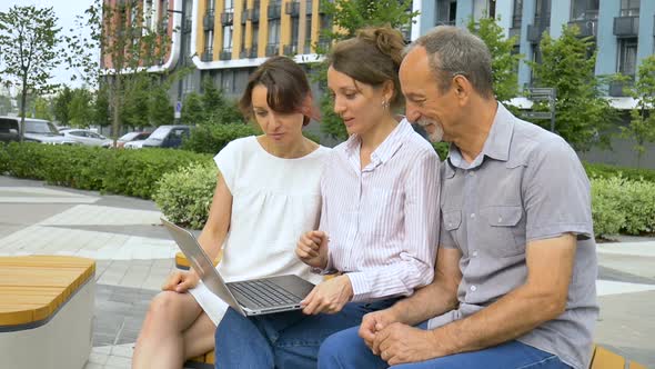 Senior Businessman and Two Female Coworkers are Discussing the Project Sitting on the Bench Near New