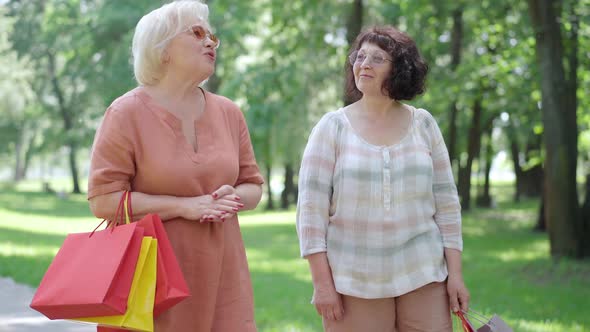 Two Positive Caucasian Senior Women Strolling in Sunny Spring or Summer Park with Shopping Bags and