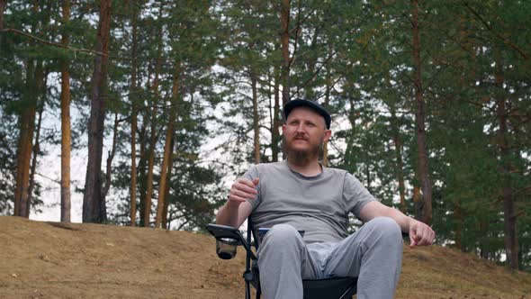 A Man with a Beard in a Cap Sits in the Woods in a Folding Chair Resting Enjoying Nature