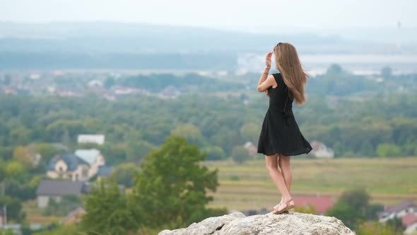 Long haired young woman in short black dress standing on hill top enjoying warm summer day.