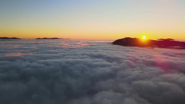 Aerial view of vibrant sunrise over white dense clouds with distant dark mountains on horizon.