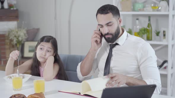 Portrait of Busy Middle Eastern Father Ignoring Little Daughter Eating Breakfast. Young Businessman