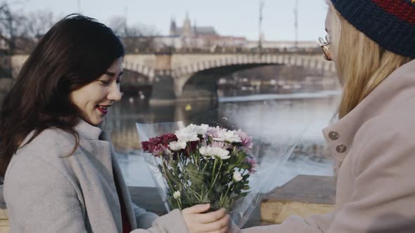 Close Friends Meeting on River Embankment and Exchanging Bouquet of Flowers in Front of Camera