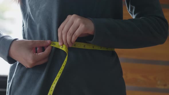 Close up Asian female measuring her waist-length with tape while standing in the living room.