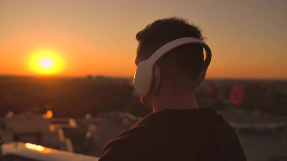 A Man Walks on the Roof at Sunset with Headphones Looking at the City From the Height of a