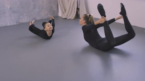 Little Asian Girl in Black Bodysuit Doing Stretching Exercises with the Help of Female Trainer