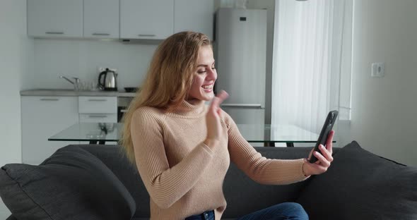 A Lovely Smiling Curly Blonde Woman is Talking to Someone with a Video Chat While Sitting in Cozy