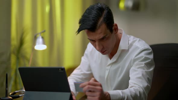 Focused Serious Middle Eastern Man Typing on Tablet Keyboard Holding Business Card in Hand