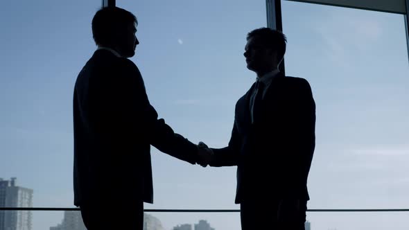 Businessmen Shake Hands And Pat Each Other On The Shoulder At Window In Office