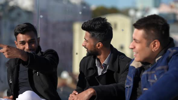 Positive Young Carefree Middle Eastern Men Talking Smiling Sitting at Glass Fence on Rooftop